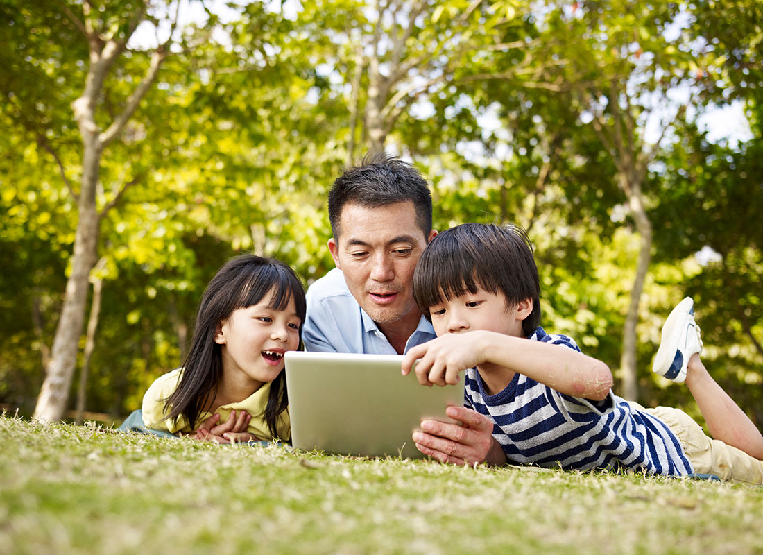 Blog - View of a Father Using a Tablet Outside with his Young Son and Daughter as They Lay on the Green Grass on a Sunny Day
